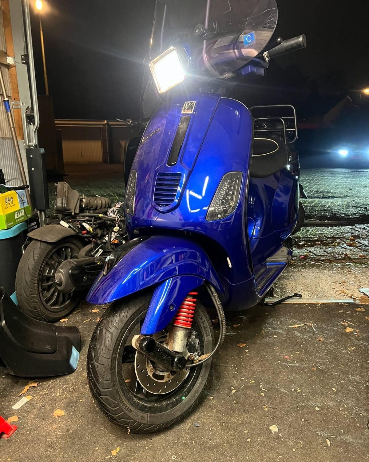 Blauwe Vespa S50 met brandende koplamp, buiten in het donker